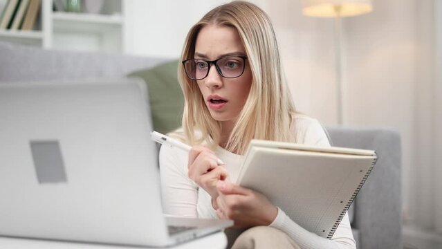Shocked focused young woman student with glasses listening interesting exciting online lecture with enthusiasm on laptop computer and make notes at home Distance remote education concept