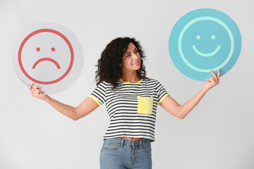Beautiful woman holding happy and sad emoticons on light background