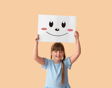 Little girl holding paper with happy emoticon on beige background