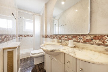 Small elongated bathroom with white porcelain sink embedded in veined marble base, frameless mirror, light tile with valances, and white shower stall