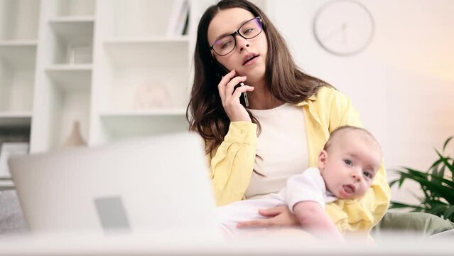 Portrait of busy young business mother holding her baby while working on laptop computer having working conversation on smartphone at light home office Successful woman balancing work and motherhood