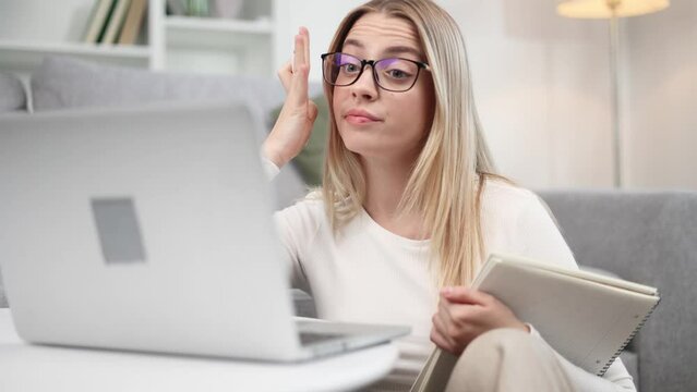 Boring unmotivated blond young woman student with glasses listening online lecture by laptop computer at light room at home Tired girl with uninteresting lesson on remote distance education indoors