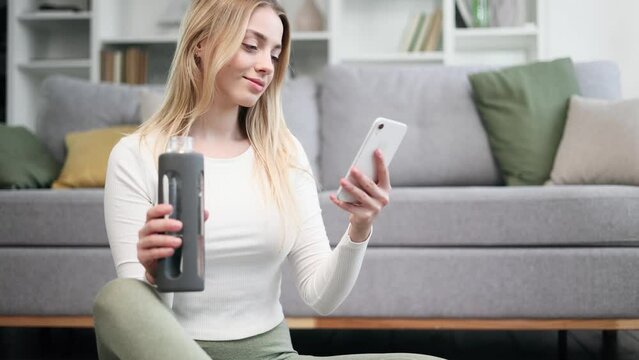 Portrait of beautiful blond woman hold smartphone watching social media with sport bottle take a break after training at home Smiling young woman texting on her phone at light room