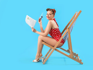 Young pin-up woman with newspaper sitting in deck chair on blue background