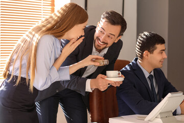 Young man and woman sprinkling pepper into their colleague's cup of coffee in office. April Fools' Day celebration