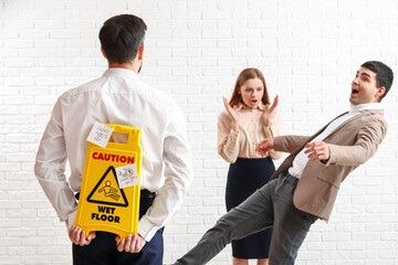Young man with caution sign playing a prank on his colleagues against white brick background. April...