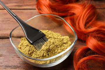 Bowl of henna powder, brush and red strand on wooden table, closeup. Natural hair coloring