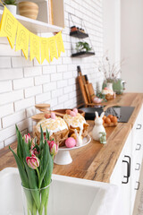 Vase with tulip flowers, Easter cakes and bunnies on kitchen counter near white brick wall