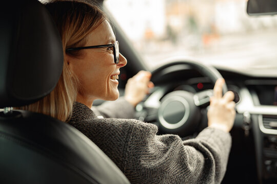 Rear view of smiling woman driving car and holding both hands on steering wheel on the way to work