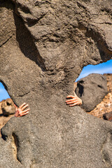 Hands only reach around the edges of a large natural stone monument sitting on a desert hillside surrounded by other scattered rocks on clear day with blue sky.