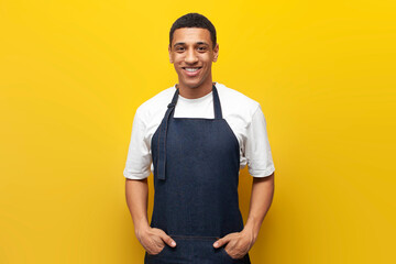 guy afro american waiter in apron on a yellow isolated background, barista worker in uniform, portrait of service man