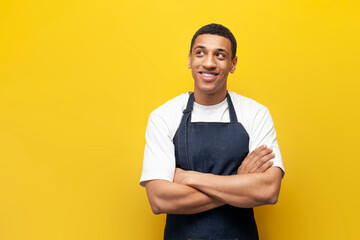 young guy afro american waiter in apron stands with his arms crossed on yellow isolated background,...