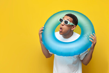 shocked african american guy in sunglasses with inflatable swim ring is surprised on yellow...