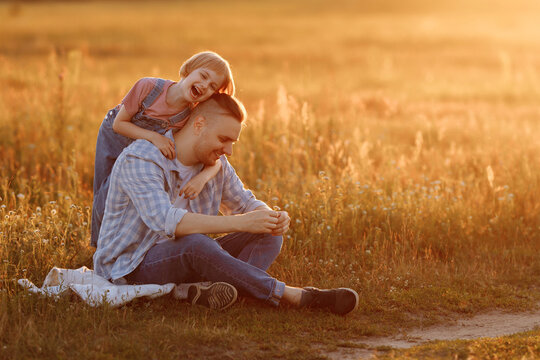 A heartwarming moment captured in time as a little girl in denim overalls embraces her father from behind while they enjoy a summer evening walk in the fields.