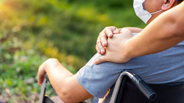 Disability Man Sit On Wheelchair  In Public Park With Sunlight.His Hand Holding By Caregiver Or Caretaker.Wheelchair For Orthopedic Patient.Medical And Insurance Concept.Copy Space With Green Trees.