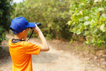 Asian boy wears blue cap, orange shirt, holds binocular in forest. Concept, Exploration. Adventure...