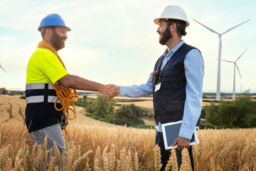 Happy Male electric engineer and smiling maintenance worker shaking hands in wind turbine farm.