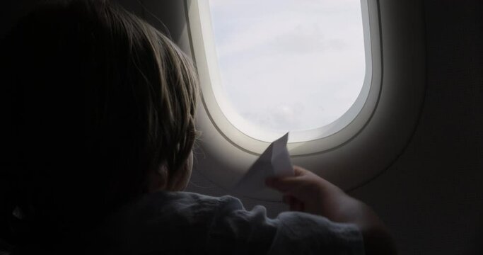 Little cute child is playing with paper plane while sitting by the window on board the aircraft. A boring and long flight on an airplane, which the boy brightens up with dreams of working in aviation.