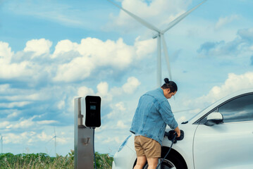 Fototapeta Progressive man with his electric car, EV car recharging energy from charging station on green field with wind turbine as concept of future sustainable energy. Electric vehicle with energy generator. obraz