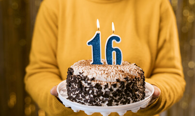 Woman holding birthday cake with burning candles number 16 against decorated background, close up....