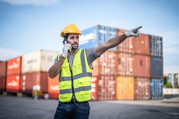 Caucasian warehouse worker in uniform with hard hat using mobile phone standing in container port terminal. Area logistics import export and shipping.