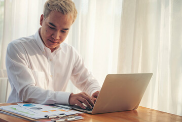 Asian man freelance using laptop at home office desk. man reading financial graph chart Planning analyzing computer data. Asian male people working office business stuff, coffee cup at home office