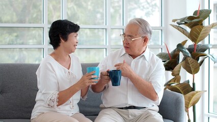 Happy Asian senior old couple sitting on sofa in living room talking enjoying tea at home, smiling middle aged mature family chatting laughing drinking coffee having fun pleasant conversation together