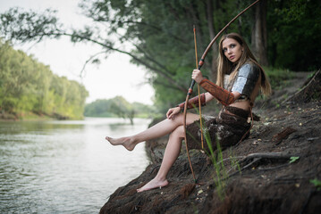 Beautiful girl warrior with the bow sits on the river shore.