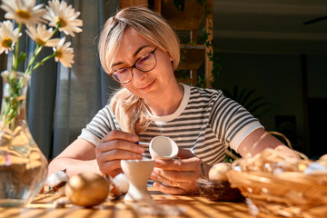 Blond woman preparing easter decoration at home, painting colorful easter eggs and coloring egg cups.