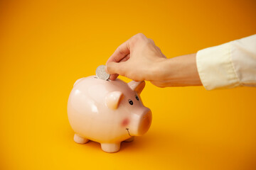 Hand putting coin into piggy bank on orange background, closeup. 
Saving or accumulation of money, investment. The concept of banking or business services.