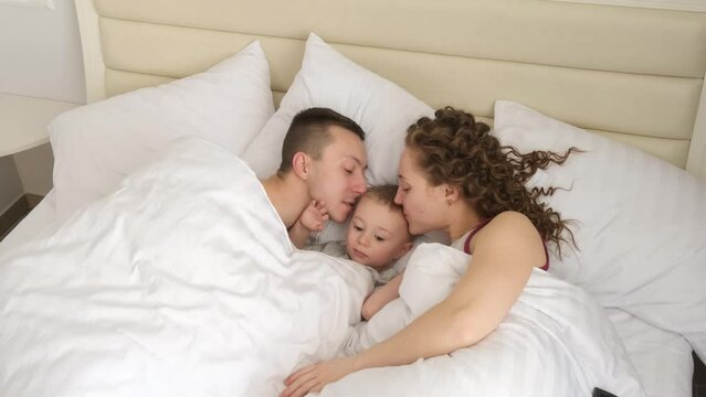 Happy young family with a child lying on the bed at home during vacation. Family relationships and family vacation concept