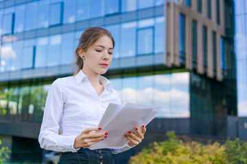 Elegant caucasian business woman in formal wear holds paper documents in her hands on the background of an office office building, an office worker near the business center.