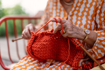 Close-up of senior woman sitting outdoor and knitting red scarf.