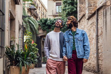 Fototapeta na wymiar gay male couple walking happy holding hands along a beautiful ancient street decorated with plants, concept of leisure and love between people of the same sex