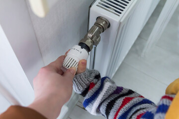 A small child with his mother warmly dressed in a sweater and a hat turns the faucet on the radiator. The concept of crisis and lack of heating in homes.