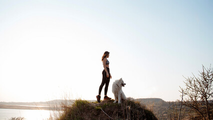 Fitness young woman standing on hill with cute fluffy dog