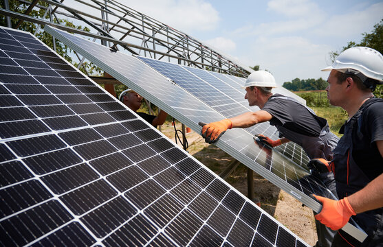 Male workers installing photovoltaic solar panel system outdoors. Men engineers placing solar module on metal rails, wearing construction helmets and work gloves. Renewable and ecological energy.