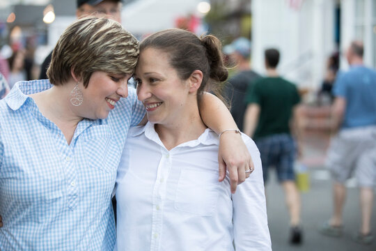 A same sex couple walks arn in arm and touches heads on a street.