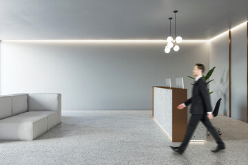 Businessman walking by modern reception desk in spacious office area with cozy grey sofa, concrete floor and light grey wall background