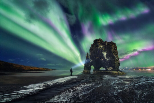 Aurora borealis above Hvitserkur rock formation, A man with a lantern (headlamp) is watching dance across night skies the Northern Lights, Hvitserkur, Golden Circle Route, Vatnsnesvegur, Iceland