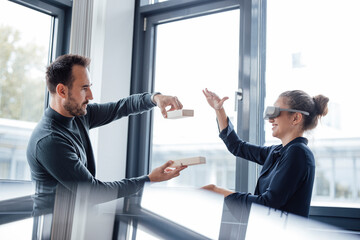 Businesswoman wearing virtual reality simulators gesturing with colleague holding wooden blocks