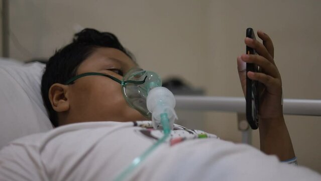 Asian 9 years old boy with oxygen mask on her face on bed in the hospital while playing cell phone