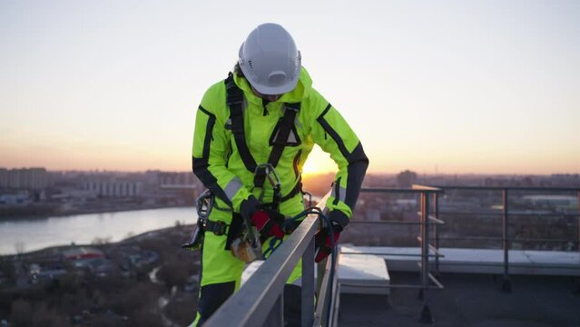 Industrial climber stands on the edge of the roof and ties a rope to descend from the building against the backdrop of the sunset