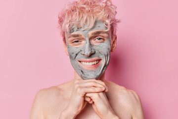 Portrait of cheerful man with rosy hair keeps hands under chin smiles happily applie nourishing clay mask stands bare shoulders has clean healthy skin isolated over pink background. Beauty procedures