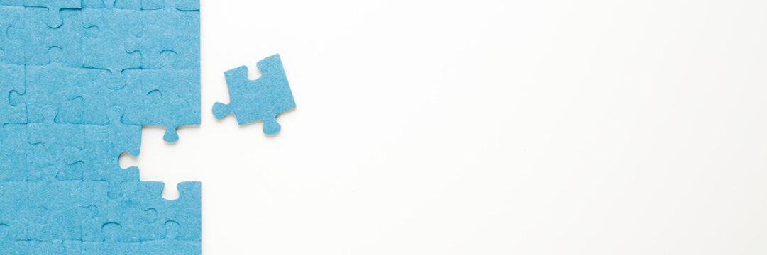 Unfinished blue puzzle with last one piece on white table background. Closeup. Wide banner. Empty place for text. Top down view.
