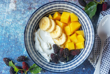 colorful yoghurt bowl with mulberry, mango and banana on blue background. yogurt bowl with fresh...