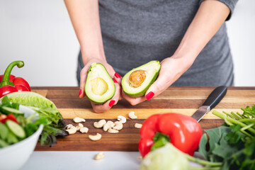 Woman with red nails holding a freshly cut avocado. Preparation of vegan salad with cashew nuts, pepper and lettuce. Raw food recipe.