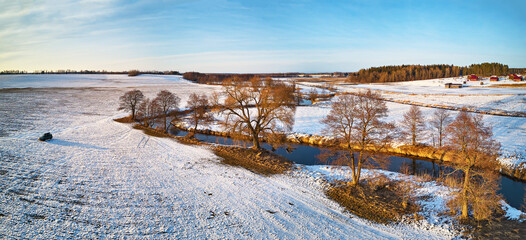 River flowing in snowy winter fields in wilderness at sunset. Stream meandering frosty countryside on cold evening. Aerial view of trees on riverbank.