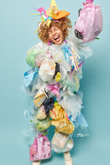 Vertical shot of curly haired female model covered with plastic garbage exclaims loudly covered collects litter poses in full length against blue background. Ecology safety and protection concept