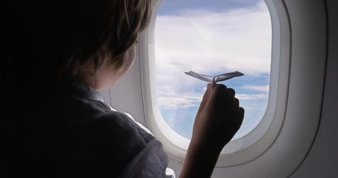 Back view, boy on plane looks dreamily out window and plays with paper plane. Child dreams of working as pilot while looking at sky and playing with the paper toy of the plane. A little future pilot.